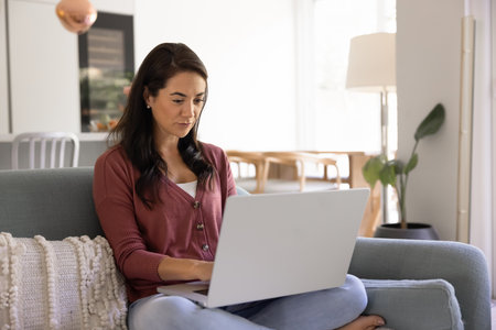 Focused young Latin homeowner woman using domestic Internet communication at home, resting with computer on sofa, typing on laptop, shopping online on e-commerce websiteの写真素材
