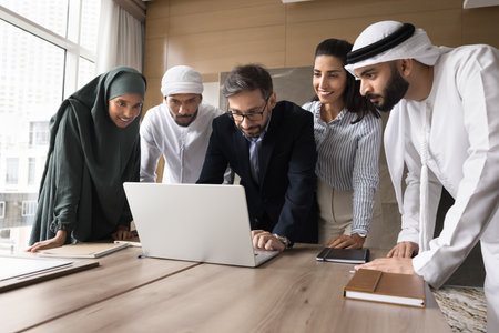 Teamwork using modern tech. Happy international team diverse young businesspeople gather around notebook at conference room study online video presentation doing research prepare electronic documentの写真素材