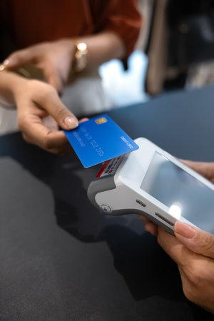 Hand of female customer holding credit card at POS terminal offered by shop assistant, making...