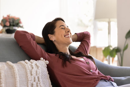 Cheerful relaxed young Hispanic homeowner woman resting on comfortable sofa with closed eyes and toothy smile, breathing cool air, laughing, enjoying indoor climate, recovery, leisureの写真素材