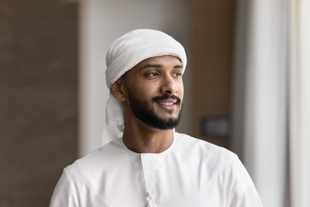 Head shot portrait handsome bearded confident young Arabian man wearing traditional Muslim white dress headwear posing indoors look aside with smile thinking dreaming creating plan idea enjoy good dayの写真素材