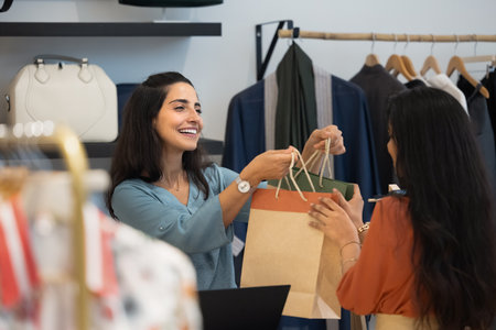 Happy Latin fashion outlet cashier giving shopping bags with purchases to young customer woman at checkout register counter, enjoying work in retail industry, connection with consumerの写真素材