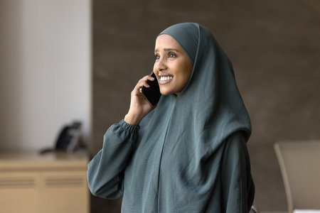Calling on business. Happy smiling young Arab girl in traditional Muslim dress talking by cellphone at modern corporate office hold pleasant conversation engaged in discussion with colleague customerの写真素材