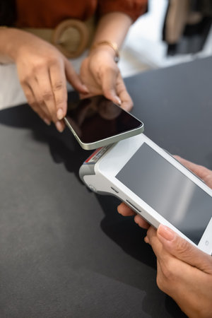 Hands of retail store customer and sales assistant holding smartphone and POS terminal with blank...