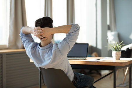 Work is done. Rear view relaxed peaceful young businessman employee recline back on office chair before laptop screen hands behind head enjoy moment of rest reduce stress after finishing project taskの写真素材