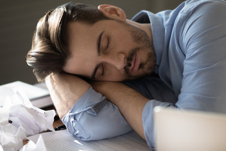 Lack of sleep. Stressed young businessman fall asleep at office workplace rest among paper balls...