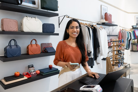 Happy young Indian fashion store manager woman posing for portrait at workplace, looking at camera, standing at cash register desk with computer and smartphone, looking at camera, smilingの写真素材