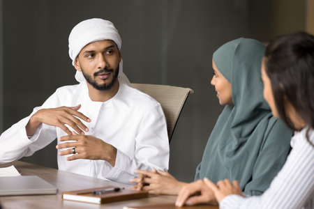 Mentoring and internship. Young Muslim male employee mentor expert in kandura consult two diverse female colleagues share professional experience explain strategy make comments after supervising workの写真素材