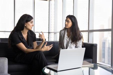 Discussing business. Confident businesswoman expert ceo project manager meet with young female colleague on couch in creative office to explain corporate marketing strategy plan using app on notebookの写真素材