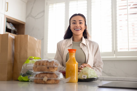 Shopping made easy and smart. Portrait happy young woman stand by kitchen table look at camera unpack fresh natural food products vegetables juice bakery received from supermarket via delivery serviceの写真素材