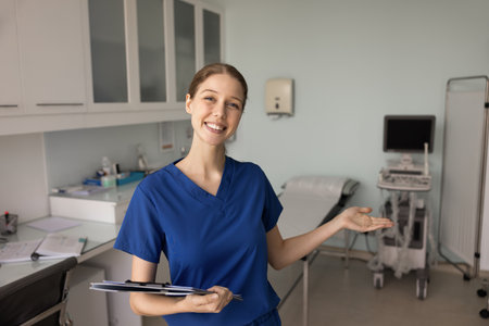 Portrait of smiling female sonographer or nurse dressed in blue scrubs holding clipboard and gesturing toward medical examination table in modern clinic room with ultrasound equipment in backgroundの写真素材
