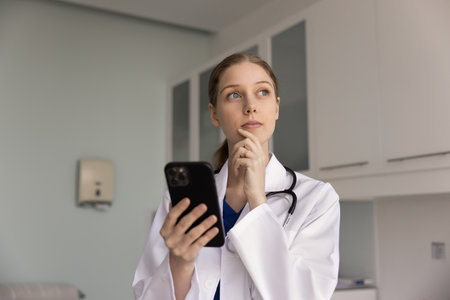Young female doctor in white lab coat with stethoscope holding smartphone and looking away thoughtfully, leading digital communication with patient, calling or reviewing client data in modern clinicの写真素材