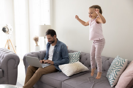 Freelance work and parenting. Smiling middle aged father remote working entrepreneur using notebook for online job sitting on comfy couch as active playful daughter preschooler jumping laughing nearbyの写真素材