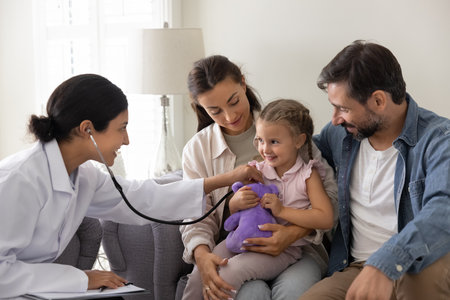Let us check your heart. Smiling young woman healthcare professional doctor nurse in white coat visiting family home check heartbeat of little patient toddler girl sitting on couch with caring mom dadの写真素材
