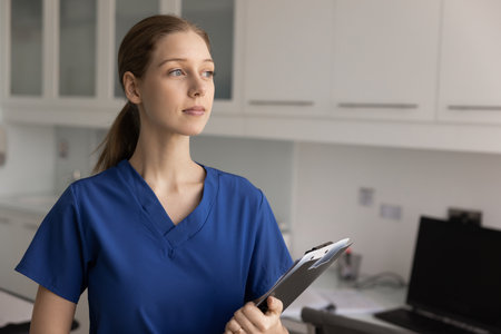 Confident female healthcare worker in blue scrubs stands in modern medical office, holding clipboard and looking thoughtfully into distance, ponders, or take break during workday at hospital. Missionの写真素材