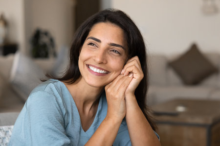 Joyful young woman posing at cozy home, sitting in living room, leaning head and cheek on hands, looking away with toothy smile, opening healthy white teeth, thinking, dreamingの写真素材