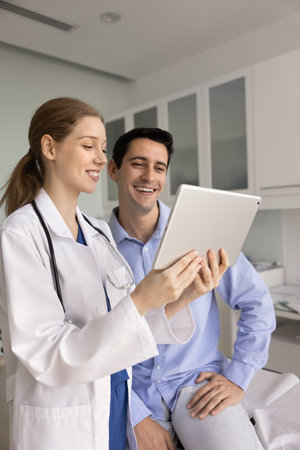 Smiling doctor and patient look at tablet screen with optimistic, joyful expression, reviewing data with good check-up health results, reading successful recovery news during professional consultationの写真素材