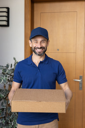 Professional courier service. Vertical portrait happy delivery man dressed in company uniform and cap posing at client home door holding cardboard box with postal parcel looking at camera with smileの写真素材