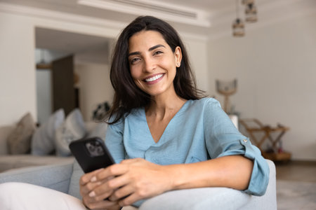 Happy beautiful Hispanic woman holding smartphone, resting in armchair at cozy home, looking at...