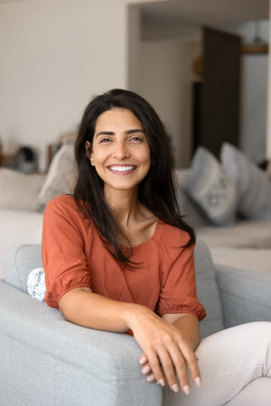 Positive beautiful young Hispanic woman home head shot. Happy Latin lady sitting in comfortable armchair, looking at camera with toothy smile, laughing, posing for vertical portraitの写真素材