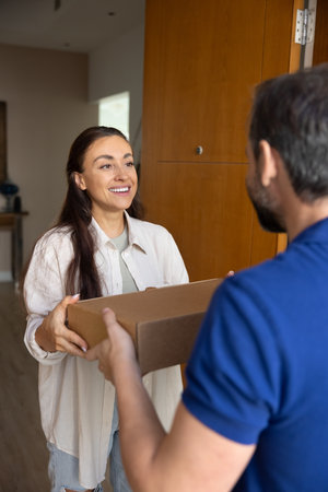 Receiving package with smile. Happy female loyal client of courier service company stand at door...