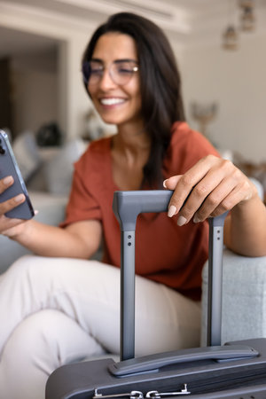 Happy young Hispanic female tourist using airlines application for online check-in, touching suitcase in hotel room, typing on mobile phone, booking apartment for vacation from home. Vertical shotの写真素材