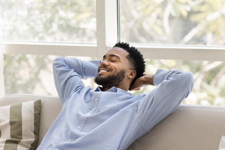 Young African man reclining on comfortable sofa with peaceful expression, enjoy pleasant, carefree rest on weekend in relaxed and serene atmosphere, relish moment of calm and tranquility at smart homeの写真素材