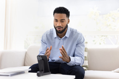 Focused African man sit on sofa, interacting with smartphone placed on stand, engaged in formal conversation via video call application, record new video for channel, share ideas or skills. Vloggingの写真素材