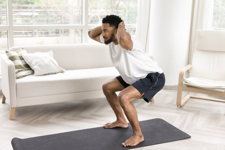 Young African man performing bodyweight squats with hands behind his head, work-out to strengthen...