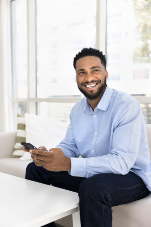 Laughing African man sits on couch with modern smartphone, enjoy new innovative platform usage, reading pleasant message, chatting with friends and family through mobile apps. Wireless internet userの写真素材