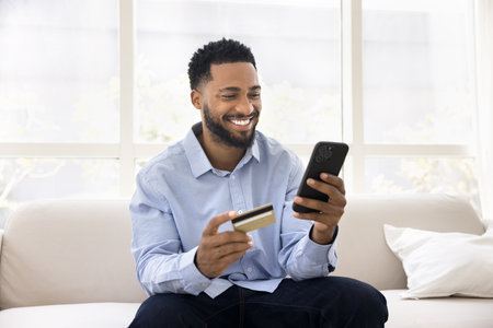 Happy African man sit on sofa with credit card and mobile phone, smile, enjoy online shopping,...