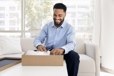 Happy African guy unpacking received gift on his personal holiday, opening package with joyful expression, exudes satisfaction and happiness from positive shopping, e-commerce or deliveries experienceの写真素材