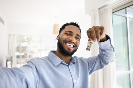 Head shot portrait happy African guy take selfie picture, making video call on smartphone camera, holding bunch of keys from his new flat, rented or purchased dwelling. First house, bank loan, tenancyの写真素材