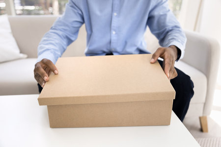Close up cropped view unknown man sits on sofa at home opens cardboard box, receiving deliveries, related to e-commerce or personal shopping, send gift to friend using reliable transportation servicesの写真素材