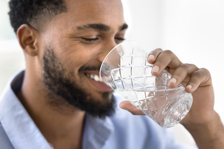 Close up view of handsome African man holds glass and drinking water, smile, make sips, enjoying...