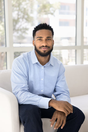 Handsome man dressed in casual blue shirt and pants sitting on couch looking at camera. African single guy spending free time in cozy living room, relaxing on weekend alone at own or rented apartmentの写真素材