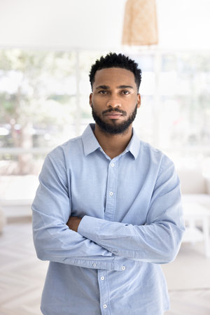 Vertical portrait handsome self-assured African man dressed in casual blue shirt posing with arms-crossed looking at camera alone, standing in living room, exuding confidence, independence and successの写真素材