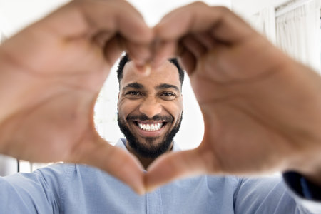 Close up African man joyfully making heart shape with hands, smiling towards camera, revealing white teeth, share happiness, promoting love and affectionの写真素材