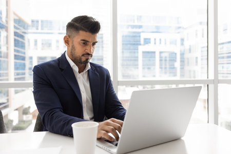 Serious young Indian executive man typing on laptop at cup of coffee, working at computer at large office window, using Internet technology for business communication. Vertical shotの写真素材