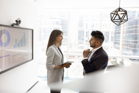Couple of young multiethnic business colleagues discussing KPI benchmarks, marketing research result in office boardroom, talking, smiling. Manager consulting analytic expert at whiteboard with chartの写真素材