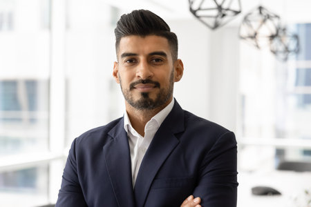 Portrait of confident young Indian business executive man looking at camera with hands folded at...