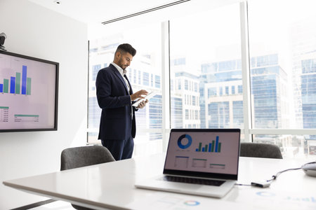 Laptop screen with chart and serious young Indian product owner analyzing Internet sales reports in background, working on marketing strategy in boardroom, typing on tablet at windowの写真素材