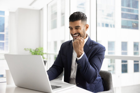 Happy overjoyed Indian businessman man celebrating online triumph at laptop, making yes winner hand...