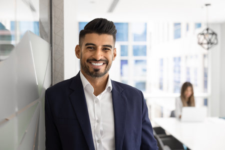 Happy handsome young Indian business leader man posing for professional portrait in office space, looking at camera, smiling, colleague working in background. CEO, executive head shotの写真素材
