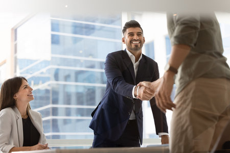 Cheerful confident Indian businessman giving handshake to investor over meeting table, getting agreement with business partner, closing deal, smiling, finishing negotiation. Low angle shotの写真素材