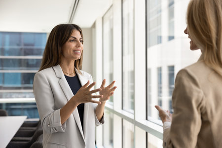Happy young Latin businesswoman talking to female business partner, standing in office meeting room, speaking to coworker woman, smiling, convincing company client, investorの写真素材
