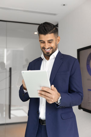 Happy young Indian businessman using online financial service on digital tablet, typing on gadget, browsing Internet, having fun, smiling, laughing, standing in office. Vertical shotの写真素材