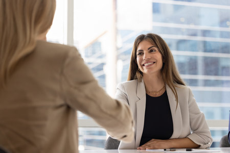 Positive young Latin businesswoman giving handshake to female job candidate after successful interview, hiring employee, getting agreement with partner, shaking hands with investor over meeting tableの写真素材