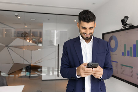 Cheerful young Indian project manager man typing on mobile phone at large office whiteboard, using Internet technology on smartphone for online communication, smilingの写真素材