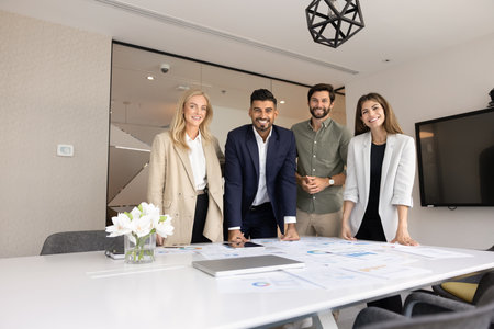 Creative team of positive young diverse professionals standing at table with heap of marketing reports, looking at camera, smiling, posing together. Group portrait of agency staffの写真素材